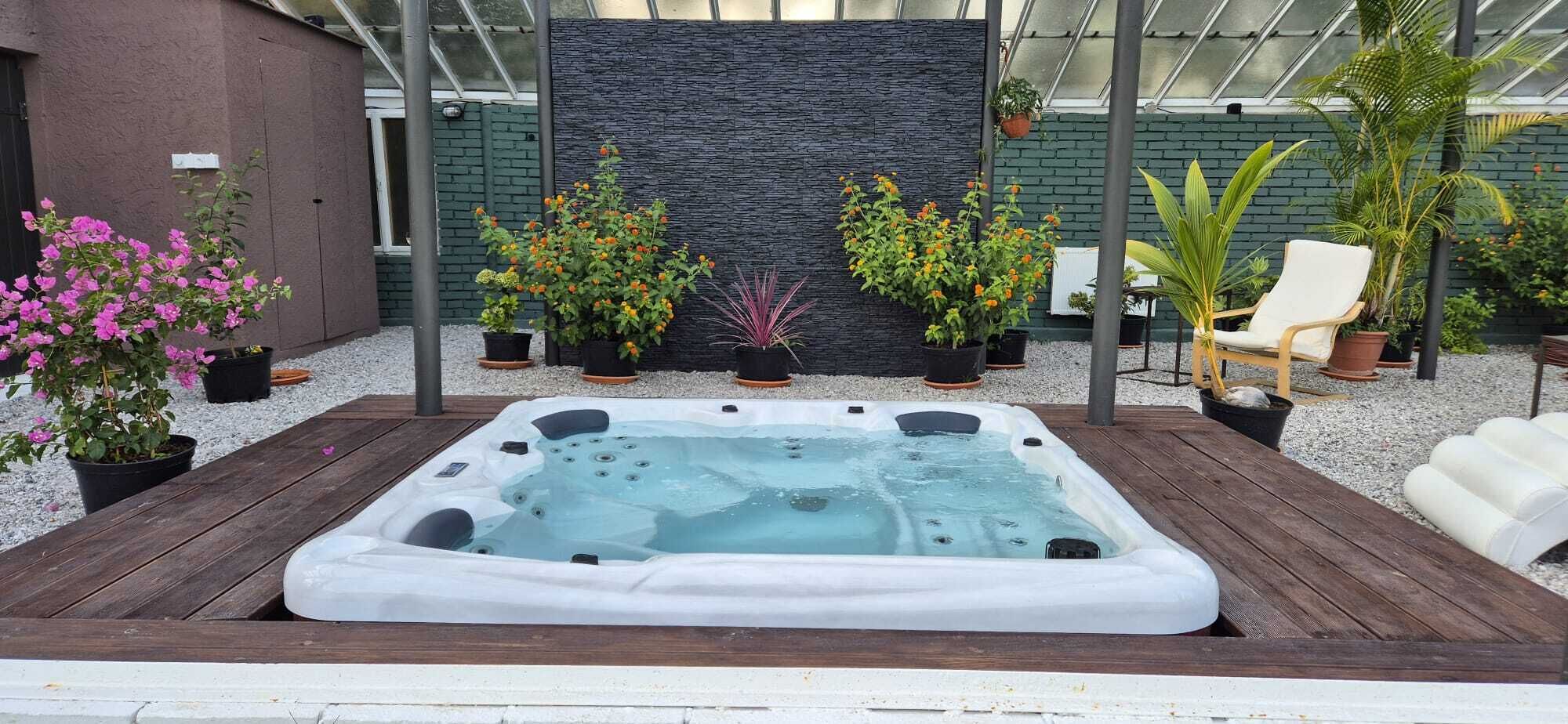 Ein Whirlpool in einer überdachten Außenbereich mit Steinwand im Hintergrund, umgeben von Topfpflanzen und Gartenmöbeln.