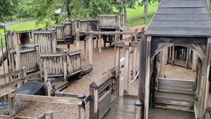 A wooden play structure with multiple levels and slides, surrounded by trees.