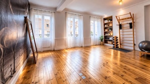 Ein geräumiger Raum mit Holzboden, großen Fenstern mit weißen Vorhängen, einer Holzleiter und einem Regal mit diversen Gegenständen.
