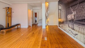 Una habitación amplia con suelo de madera, un banco, una máquina de ejercicios montada en la pared y una escalera que conduce a otra habitación.