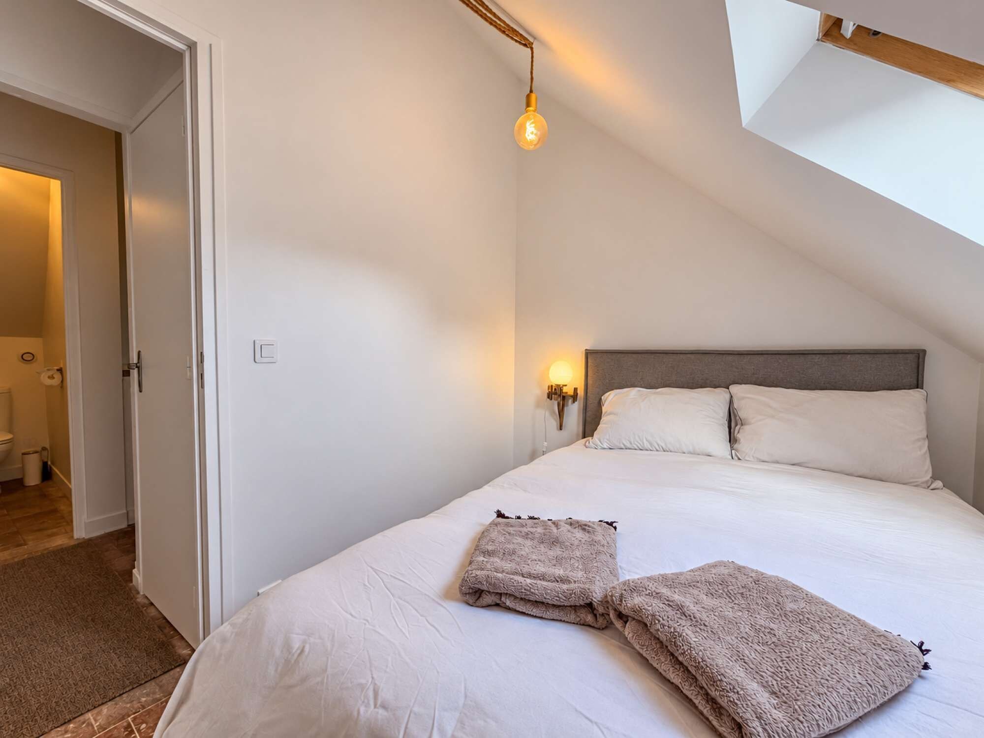 La chambre avec fenêtre de toit vue depuis l’autre angle. Le cadrage montre le lit, la sous-pente et l’ouverture vers l’espace sanitaire.