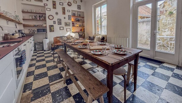 Una cocina amplia con un gran comedor listo para cuatro personas, rodeado de sillas. Hay varias estanterías con utensilios de cocina y cuadros en las paredes.