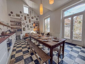 Une cuisine spacieuse avec une grande table à manger, dressée pour quatre personnes et entourée de chaises. De nombreuses étagères, remplies d’ustensiles de cuisine, et des tableaux encadrés ornent les murs.
