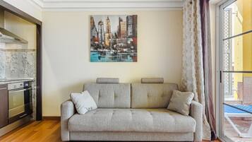 A living room with a grey sofa, a painting of a cityscape, a kitchen with a stove, and a window with a view of a pool.