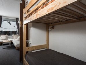 Ein Etagenbett mit Holzbettgestell und einem Bett darunter. Ein Fenster mit Vorhängen bietet Blick ins Freie. Im Raum sind ein kleiner Tisch und ein Stuhl zu sehen.