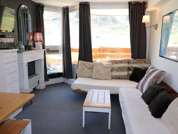 Ein gemütliches Zimmer mit einem weißen Sofa, einem kleinen Holztisch und Blick auf eine Holzterrasse durch große Fenster.