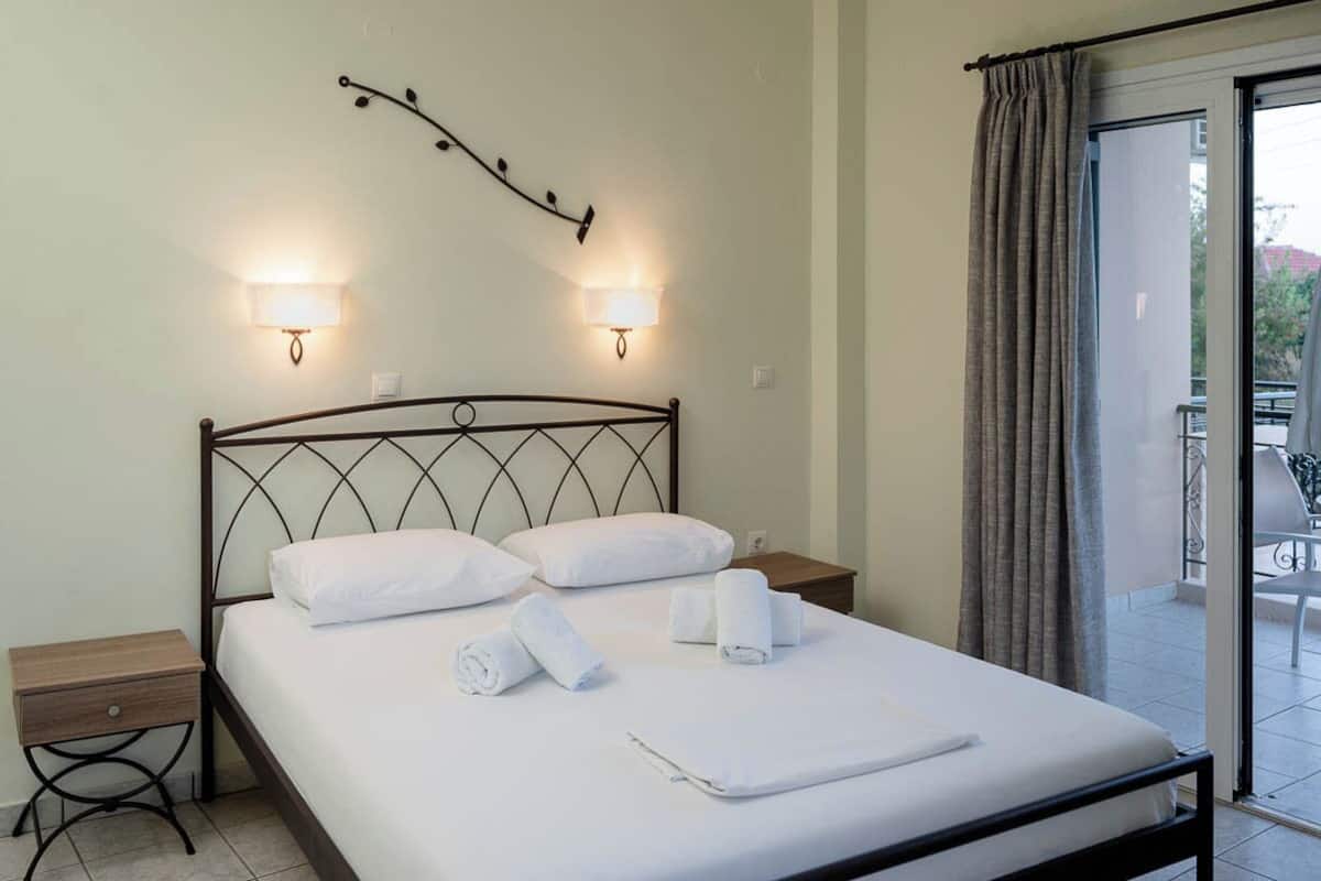 A neatly made bed with white linens and rolled towels, two wall-mounted lamps, and a balcony with a curtain.