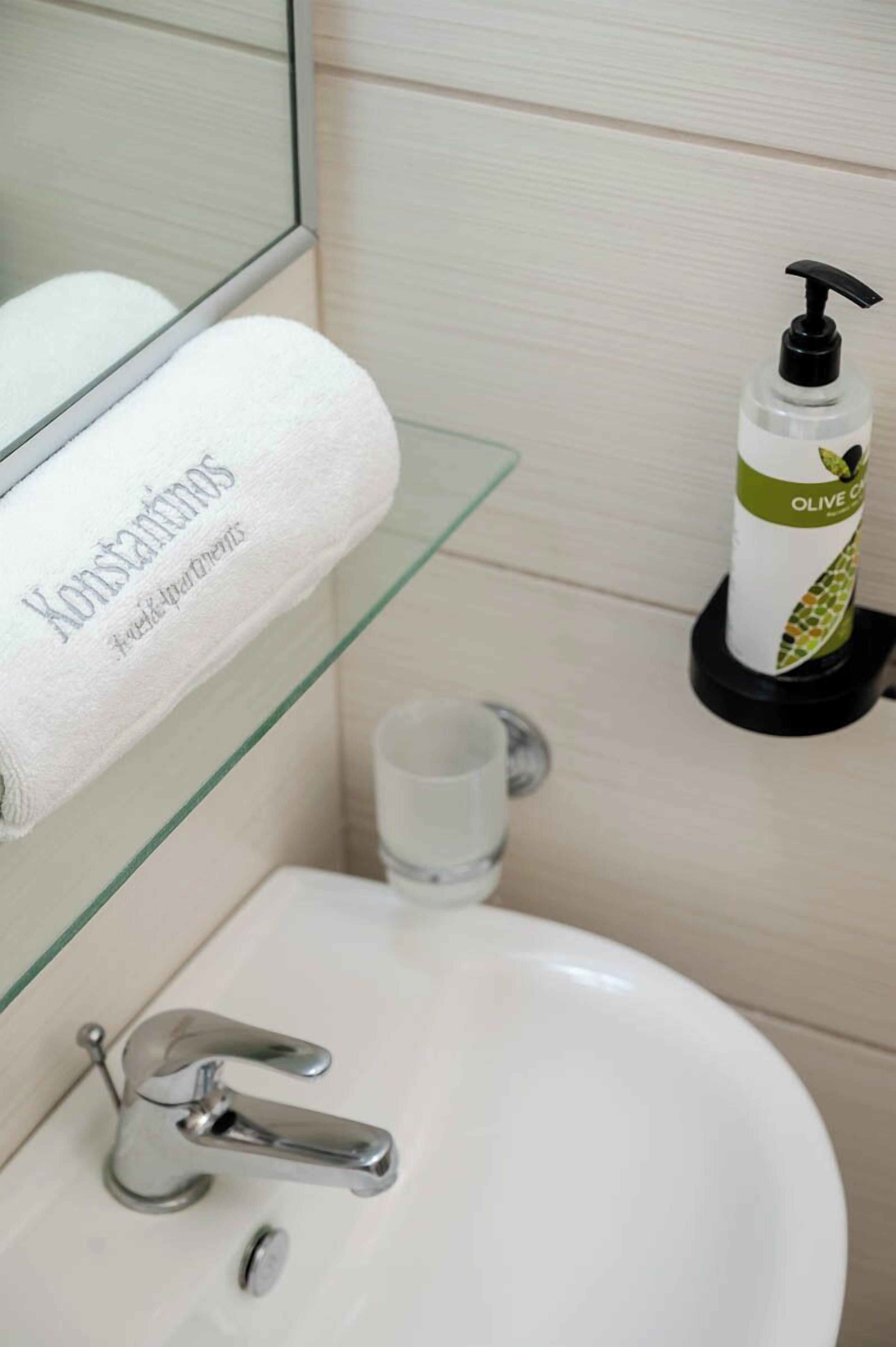 Une salle de bain avec un lavabo blanc, une commode avec un plateau en verre, une serviette blanche ornée d’un motif, et une bouteille d’huile d’olive.