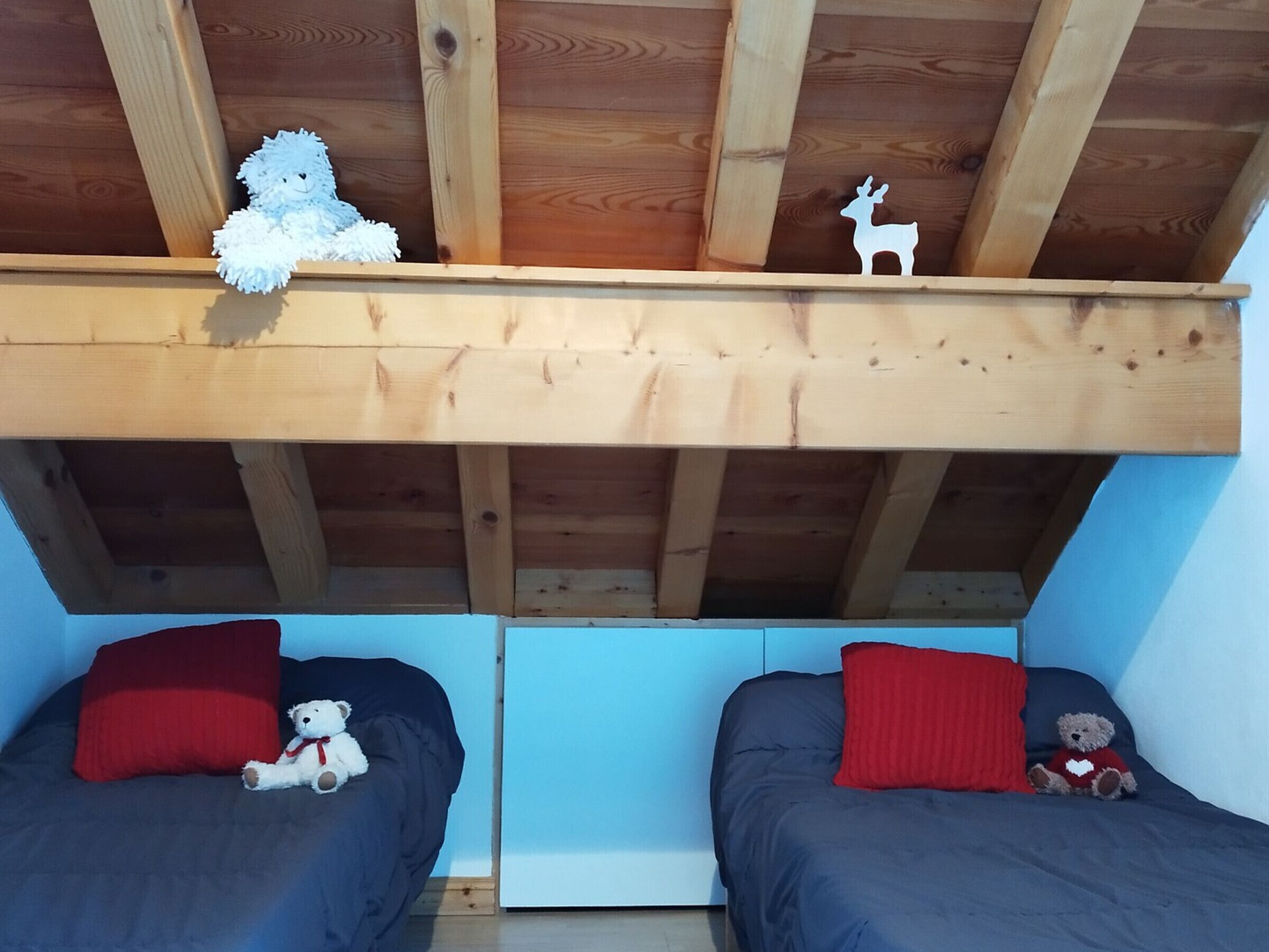 Une chambre avec deux lits, des poutres en bois et un espace en mezzanine agrémenté d’un ours en peluche et d’une découpe en forme de cerf.