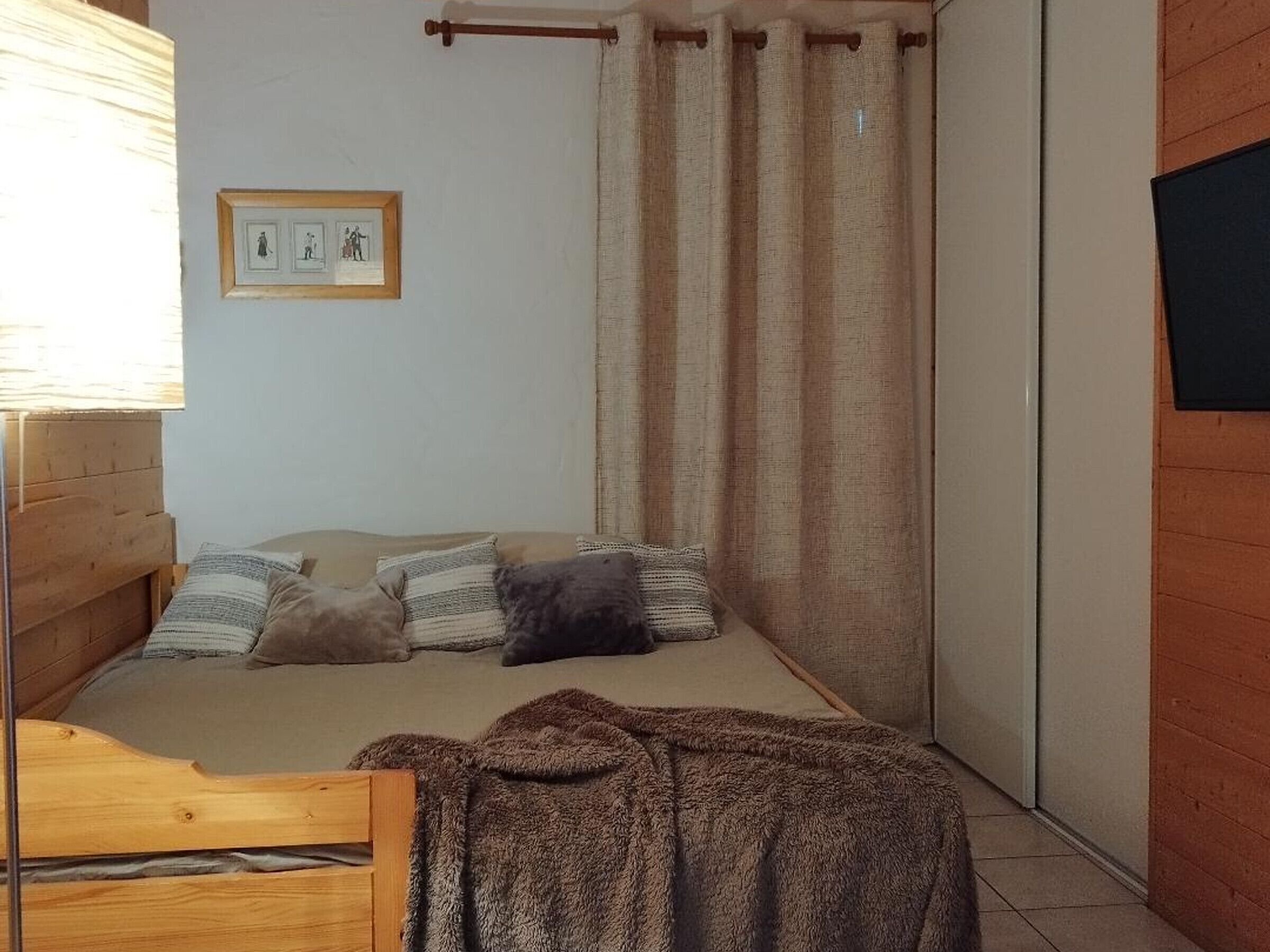 A small, cozy room with a bed, a wooden headboard, a window with blinds, a curtain, and a framed picture on the wall.