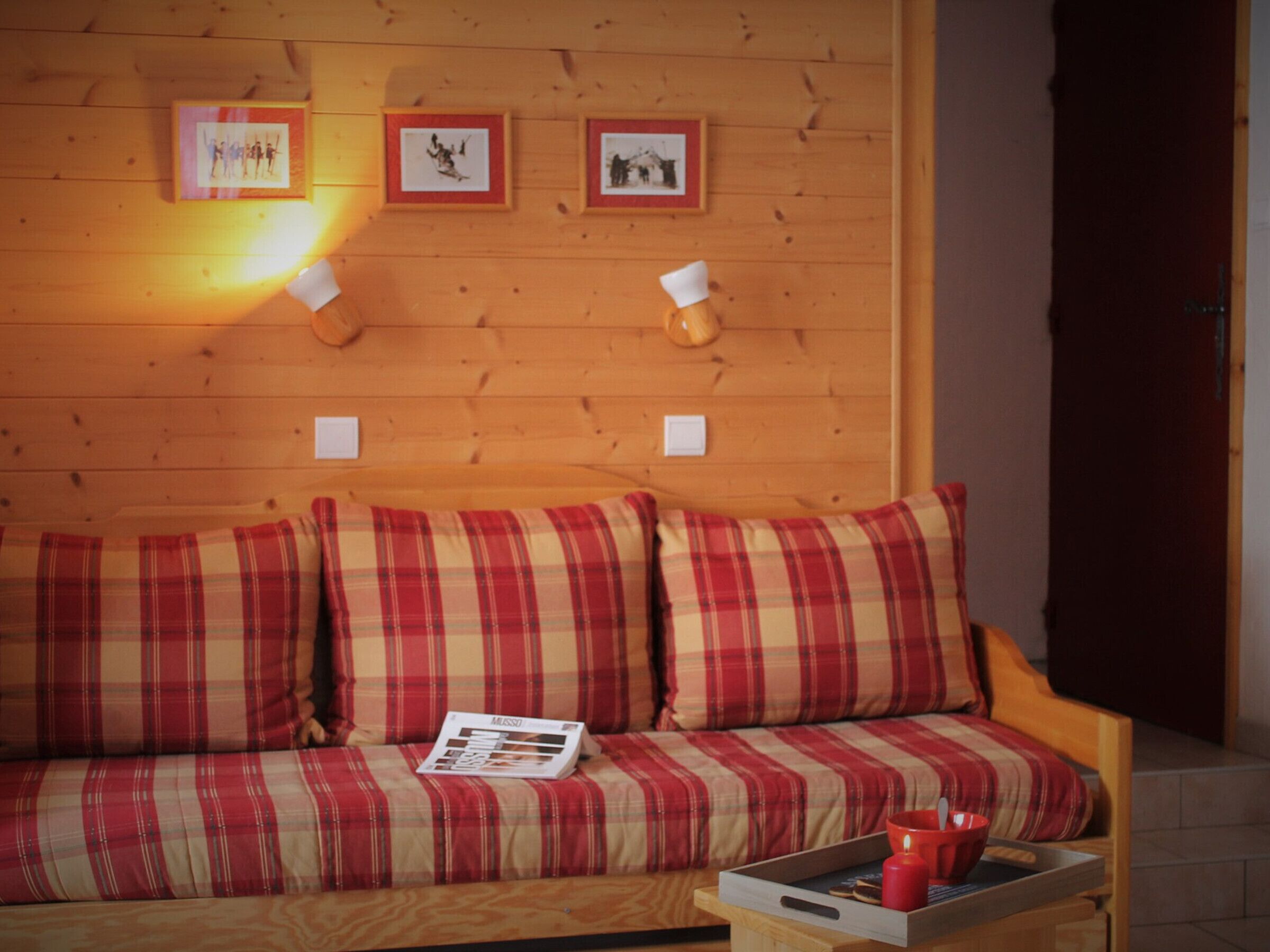 Pared de madera con cuadros enmarcados, sofá escocés con una revista, y una bandeja con galletas y una vela.