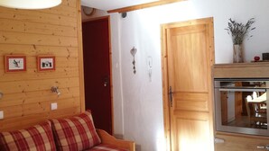 Quarto aconchegante com parede de madeira, porta vermelha, sofá com almofadas xadrez e cozinha com fogão.