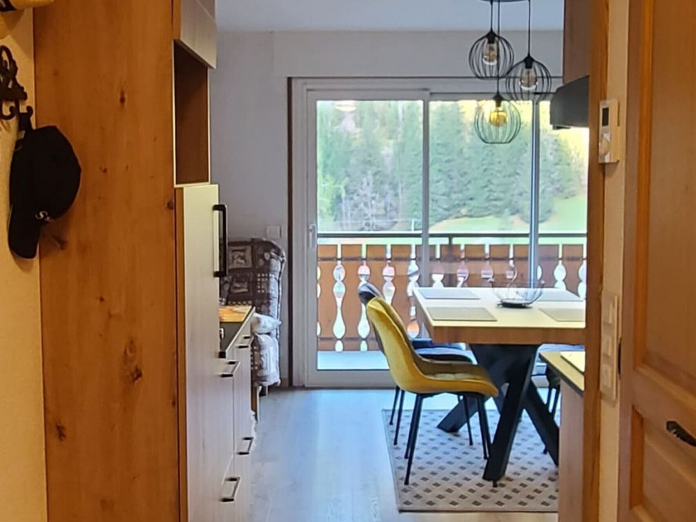 A room with a wooden door, a dining area with a table and chairs, a view of the outdoors through a large window, and a balcony with a railing.