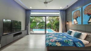 A modern bedroom with a large bed, a flat-screen TV, and a view of a swimming pool and greenery. - 3BR Near Beach Tub & Pool Table V204 (Rawai)