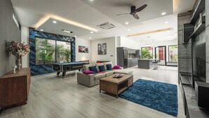 A modern living room with a sofa, coffee table, and a pool table. There is a kitchen area in the background with a large window. - 3BR Near Beach Tub & Pool Table V204 (Rawai)