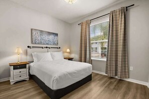 Une chambre à coucher avec un lit, des tables de chevet, un tableau, une fenêtre avec des rideaux et une vue sur l’extérieur.