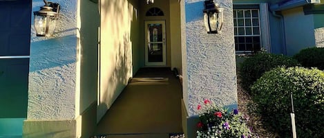 En byggnadsentré med en överbyggd gång, stenmurar och en gångväg som kantas av småsten och blommor.
