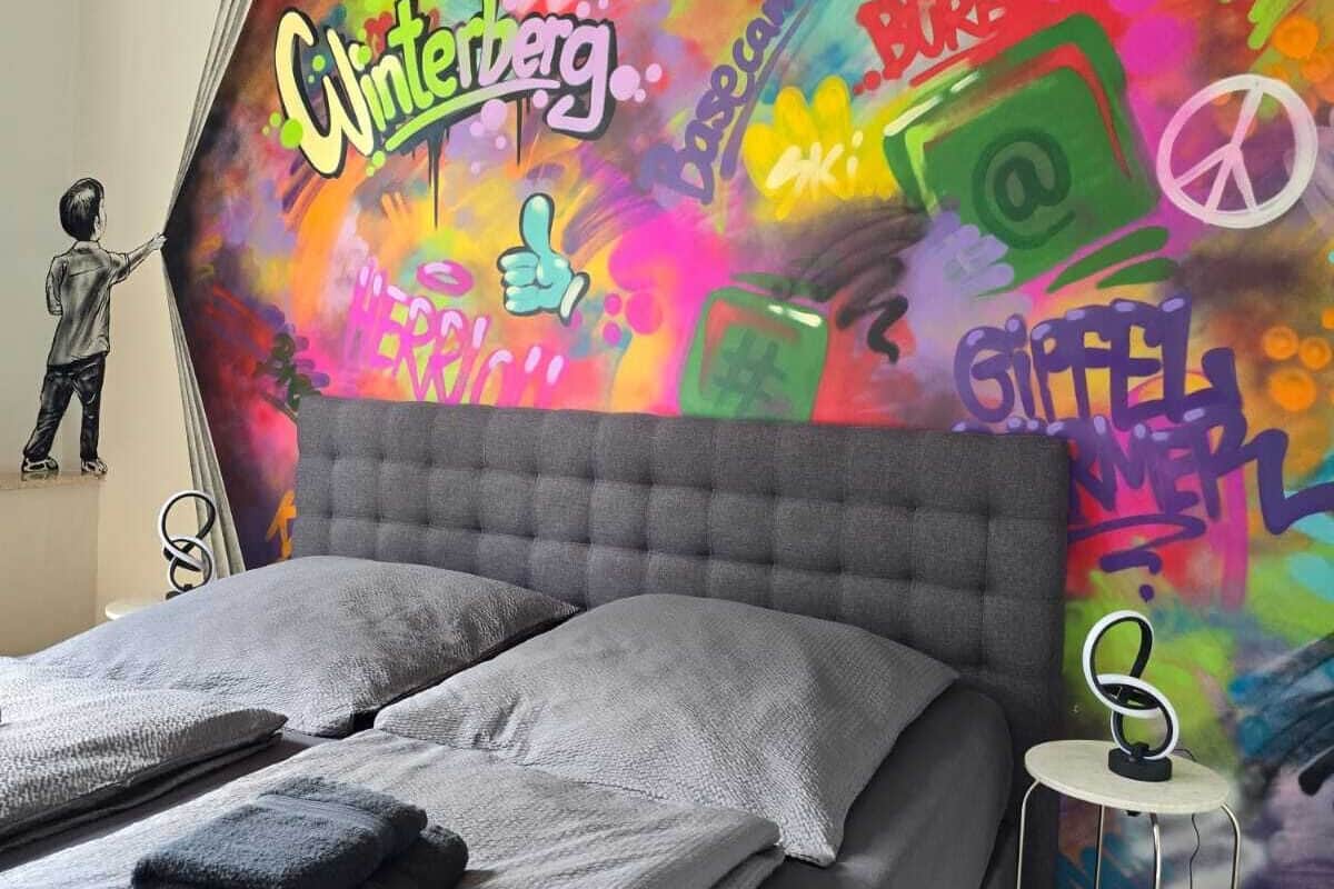 A bedroom with a colorful graffiti-style wall mural, a tufted headboard, a bed with grey linens, and a small bedside table with a lamp.