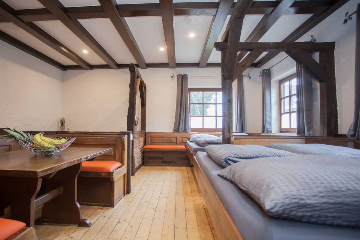 A room with wooden beams, a wooden table with a fruit bowl, a bench, and a bed with pillows.