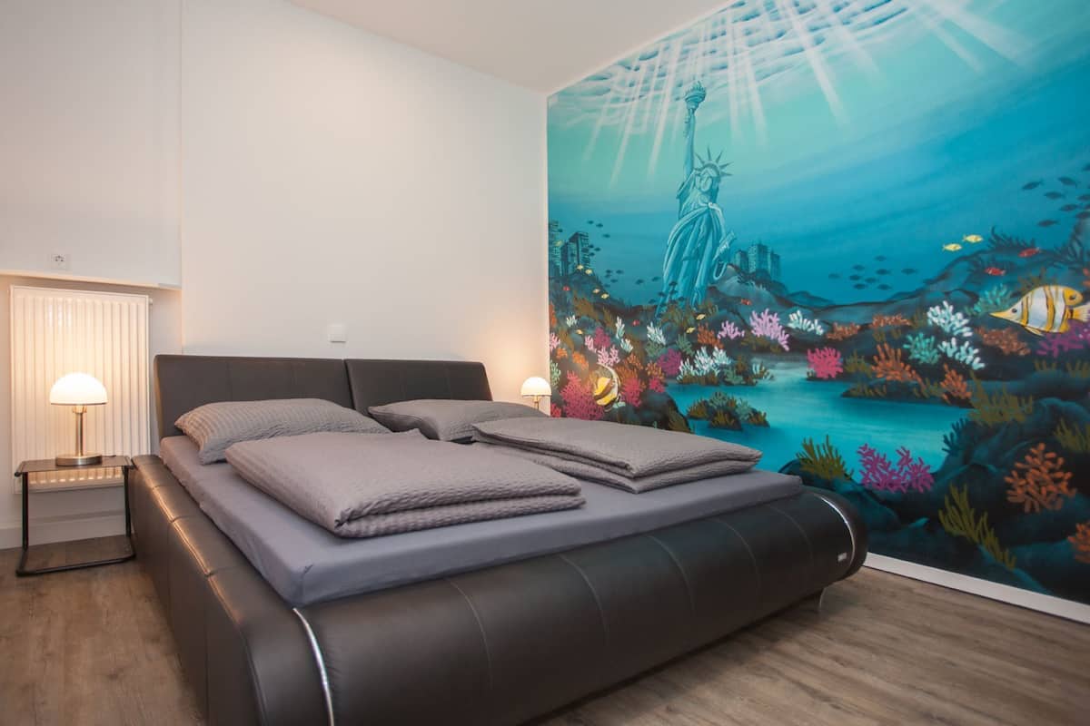A bedroom with a bed, two bedside tables, a nightstand, and a mural of an underwater scene with a statue.