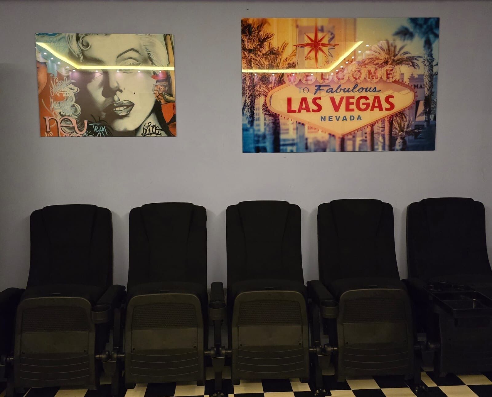 Eine Reihe schwarzer Stühle auf einem Boden mit Schachmuster, eine Wand mit zwei Plakaten, von denen eines "WELCOME TO Fabulous LAS VEGAS NEVADA" zeigt.