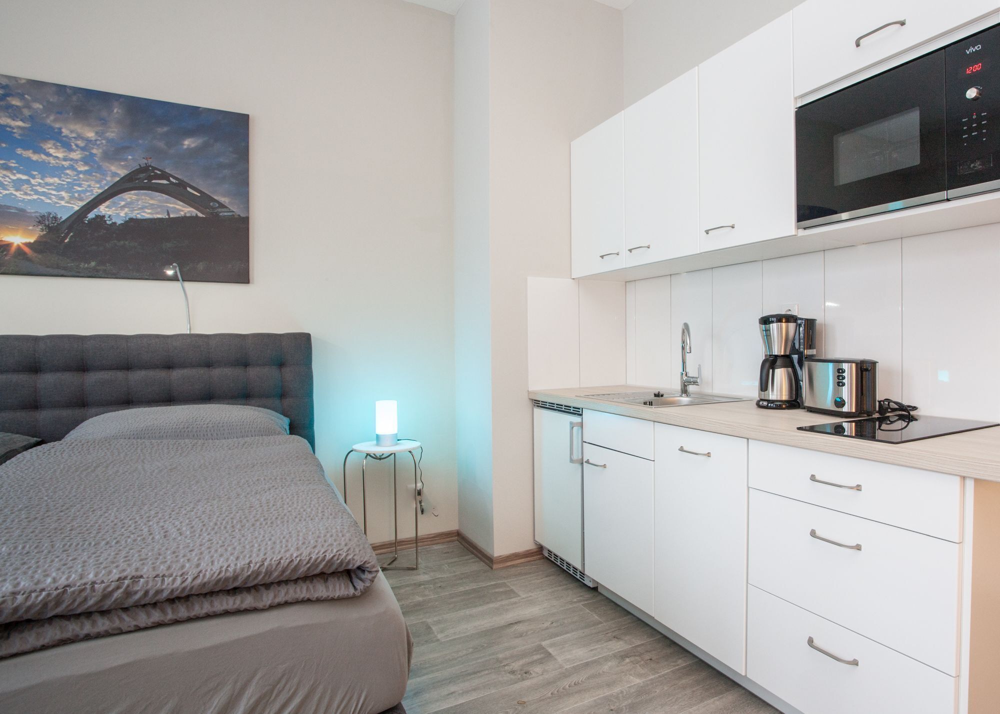 A modern hotel room with a bed, a small bedside table with a lamp, a kitchenette with a microwave, coffee maker, and toaster, and a painting on the wall.