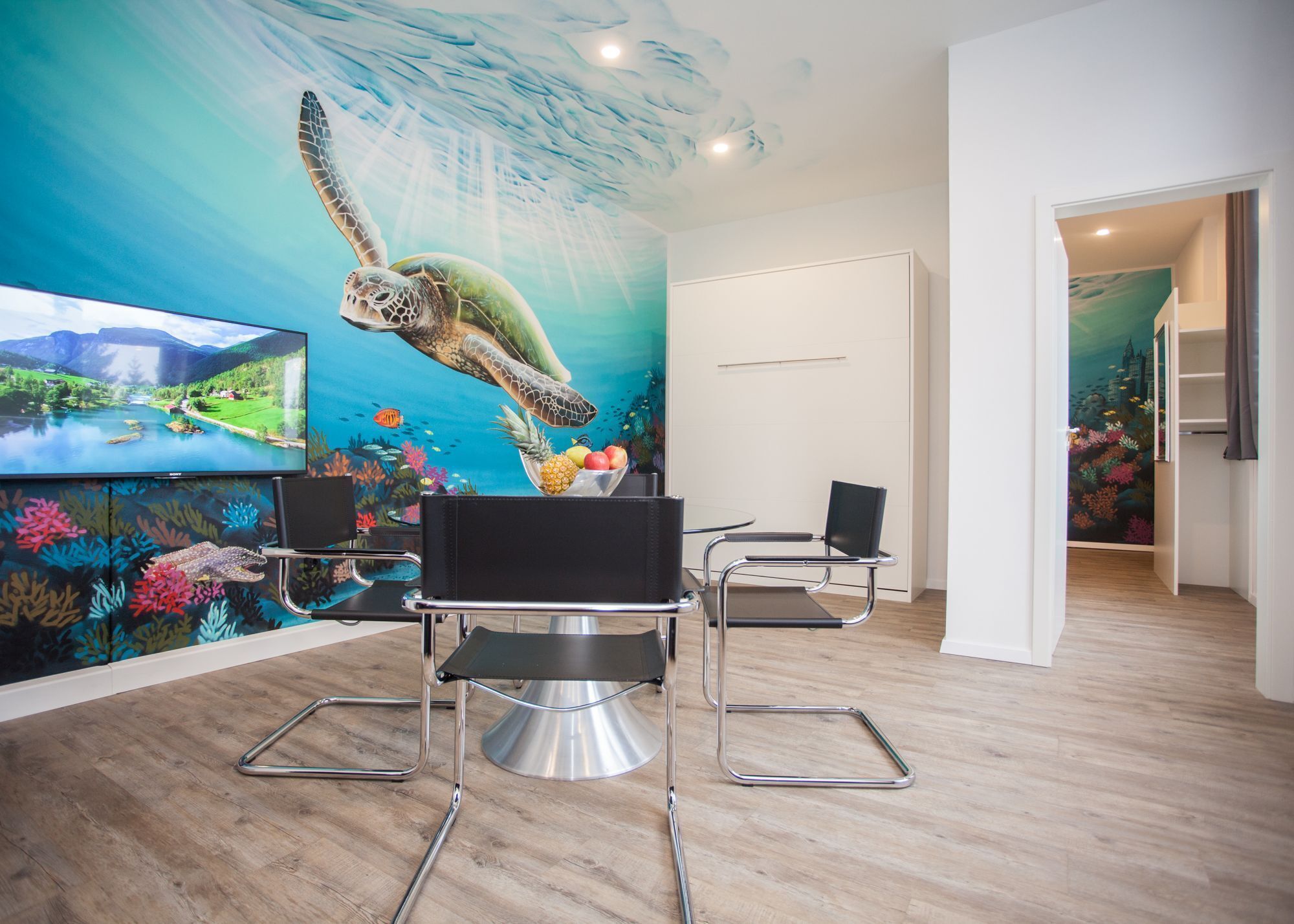 Quarto moderno com um mural de tartaruga marinha, uma TV de tela plana e área de jantar com cadeiras de metal.