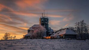 Un paysage enneigé avec un grand château d’eau, plusieurs bâtiments et des arbres sans feuilles, le tout sous un ciel au coucher du soleil.