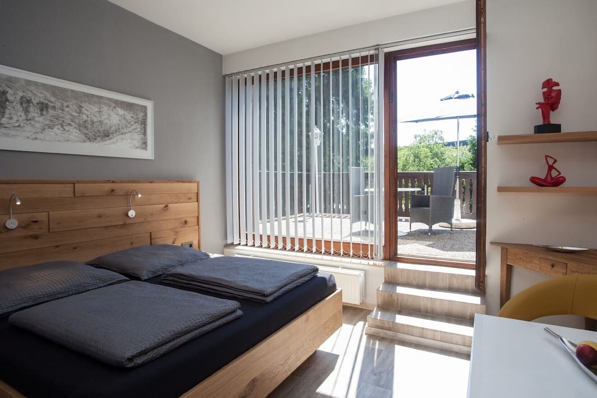A bedroom with a wooden bed, a large window with blinds, a small table, and a chair.