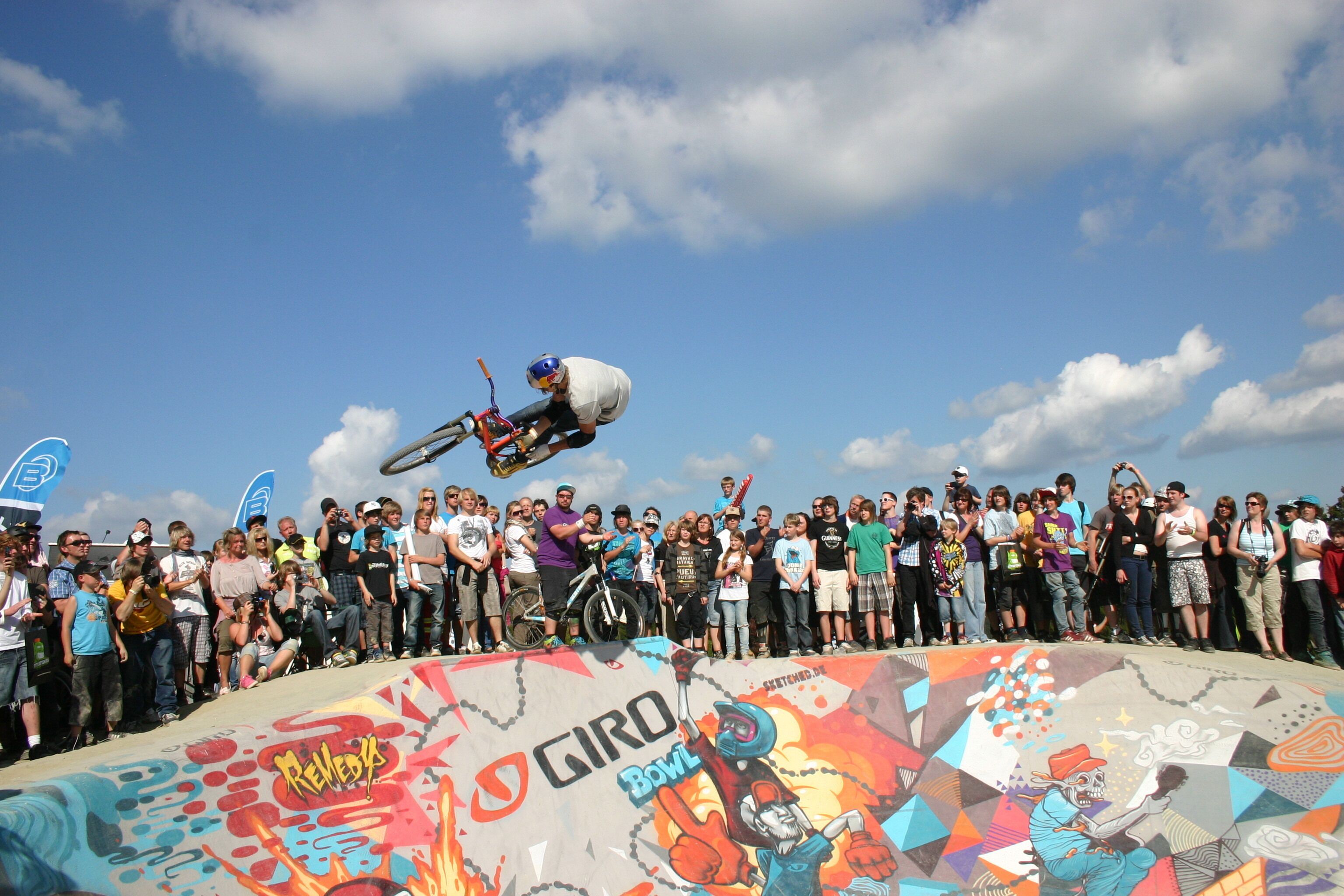 Een skateboarder voert een truc uit in een skatepark, terwijl een menigte toeschouwt.