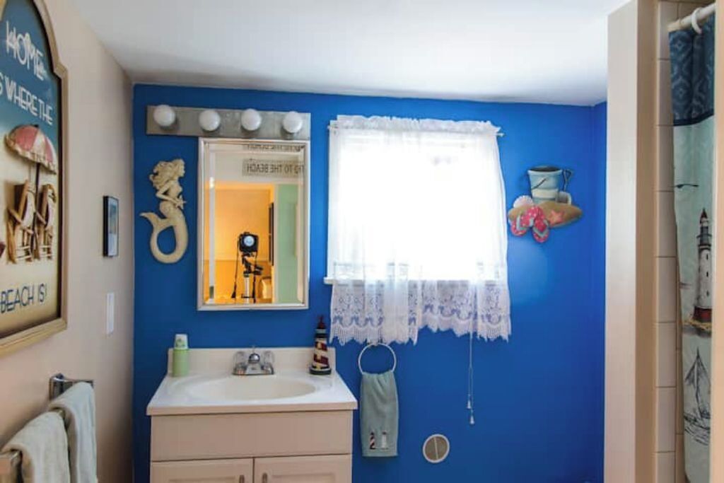 Un baño con pared azul, un lavamanos blanco, un espejo y una ventana con cortinas de encaje.