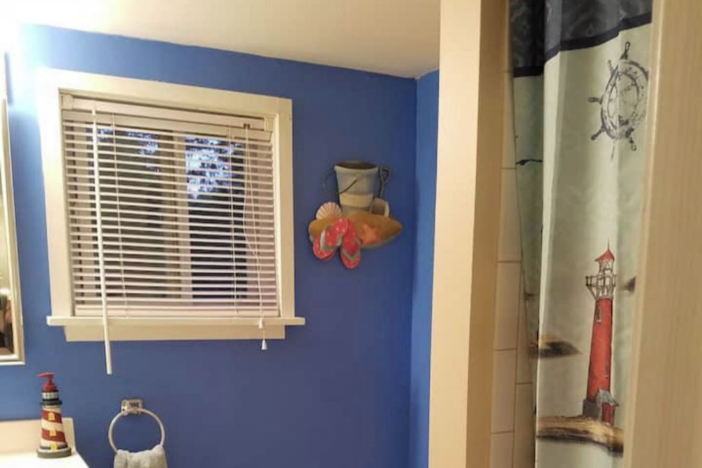 A bathroom with a blue wall, a window with blinds, a towel ring, a bottle of hand soap, and a shower curtain with a lighthouse design.