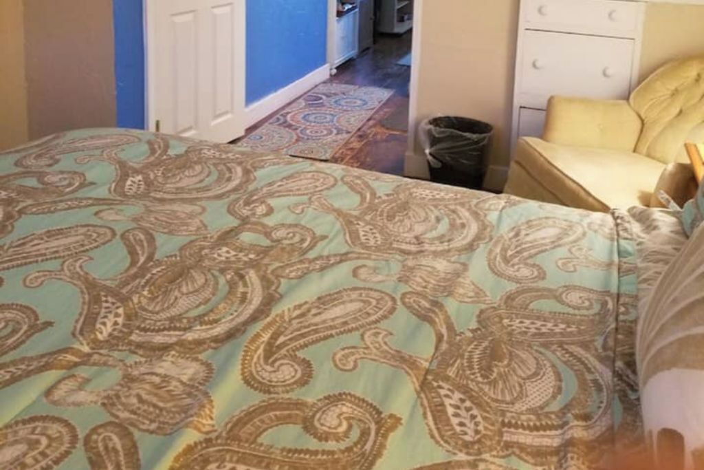 Un dormitorio con una cama cubierta por una colcha de estampado paisley, un sillón amarillo, un aparador blanco y un cubo de basura a la vista.