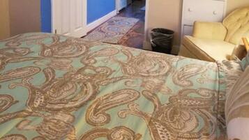 Un dormitorio con una cama cubierta por una colcha de estampado paisley, un sillón amarillo, un aparador blanco y un cubo de basura a la vista.