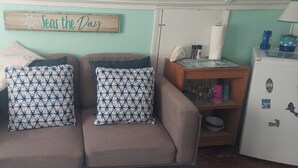 Een woonkamer met een bank, gestreepte kussens, een houten bijzettafeltje met een lamp, een koelkast, en een bord waarop staat: 