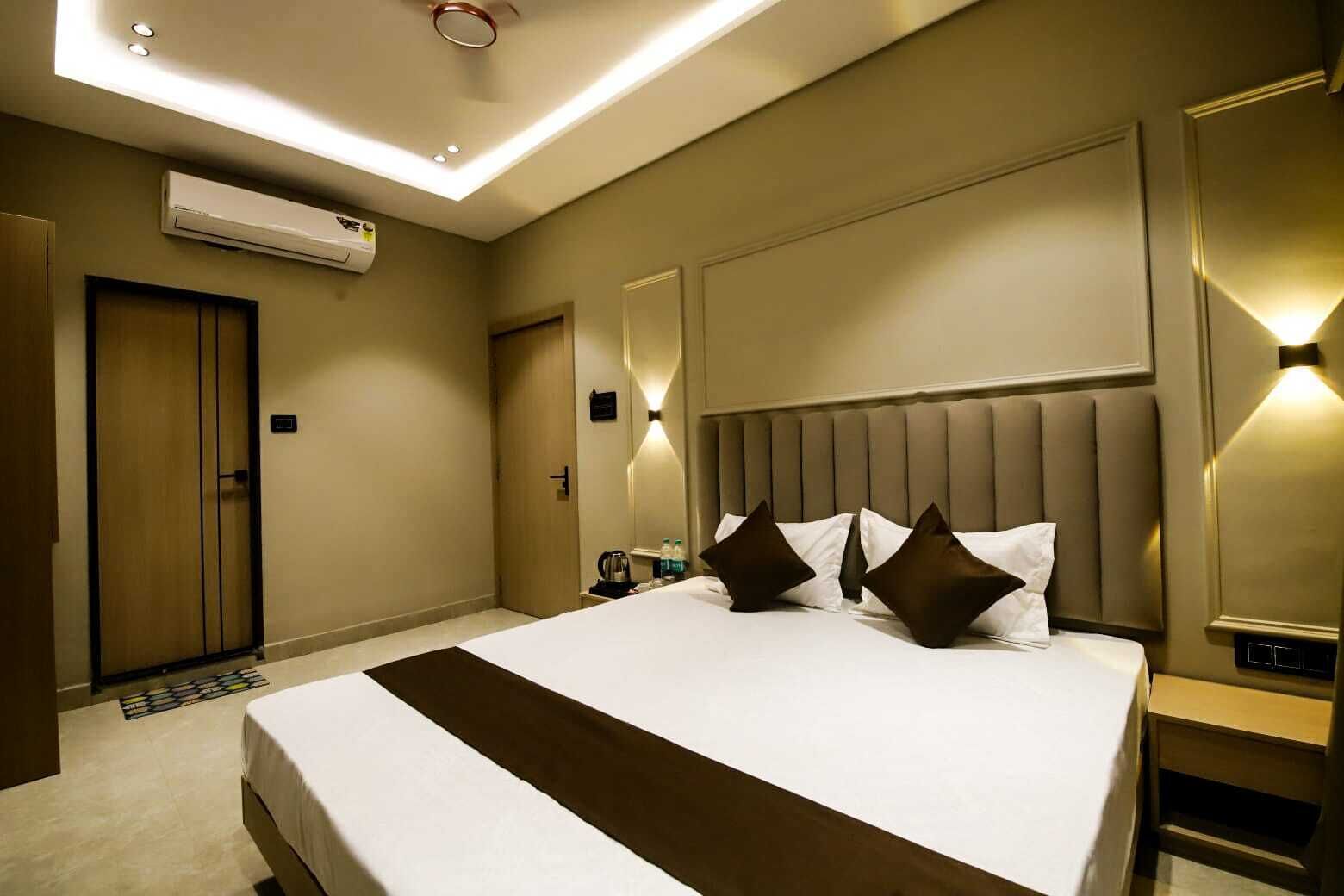 A hotel room with a large bed, a wooden door, an air conditioning unit, a bedside table with a lamp, and a small table with a kettle and a bottle.