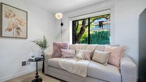 A modern living room with a grey sofa, multiple cushions, a small side table with a plant, a floor lamp, and a large window with a view of greenery and buildings.