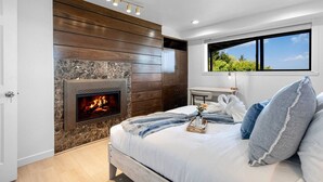 A bedroom with a stone fireplace, wooden panel wall, a bed with white bedding, and a view of greenery through a window.