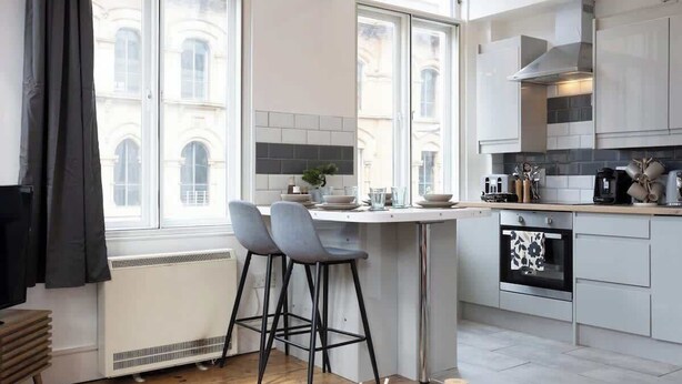 Una cocina moderna con una isla central, taburetes de bar y vista a un edificio a través de amplios ventanales.