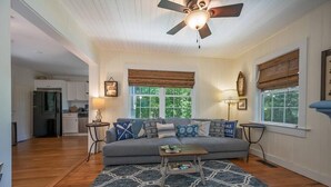 A spacious living room with a grey sofa, patterned rug, and a ceiling fan. A kitchen is visible in the background.
