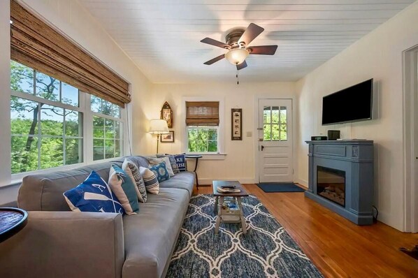 A living room with a sofa, patterned rug, ceiling fan, fireplace, and large windows. - Wish Upon the Mtn: Lake Lure + Hot Tub + Views! (Lake Lure)