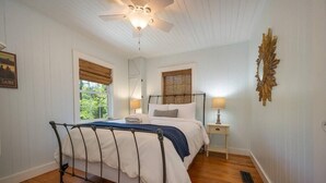 A bedroom with a bed, a ceiling fan, a window with blinds, a nightstand, and a wall-mounted mirror.