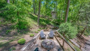 A forested area with a fire pit, two Adirondack chairs, and a wooden deck.