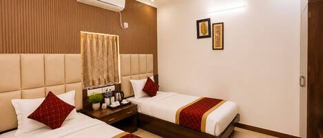 A hotel room with two beds, a ceiling fan, an air conditioning unit, a bedside table with a lamp, and a wall-mounted picture.