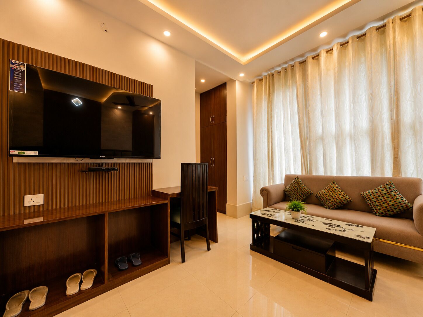 A modern living room with a flat-screen TV, a sofa with patterned cushions, a coffee table, and a shoe rack.