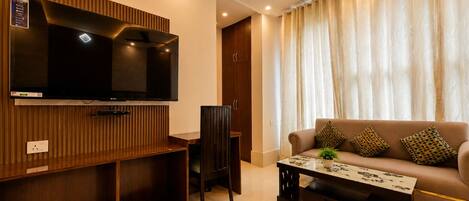 A modern living room with a flat-screen TV, a sofa with patterned cushions, a coffee table, and a shoe rack.