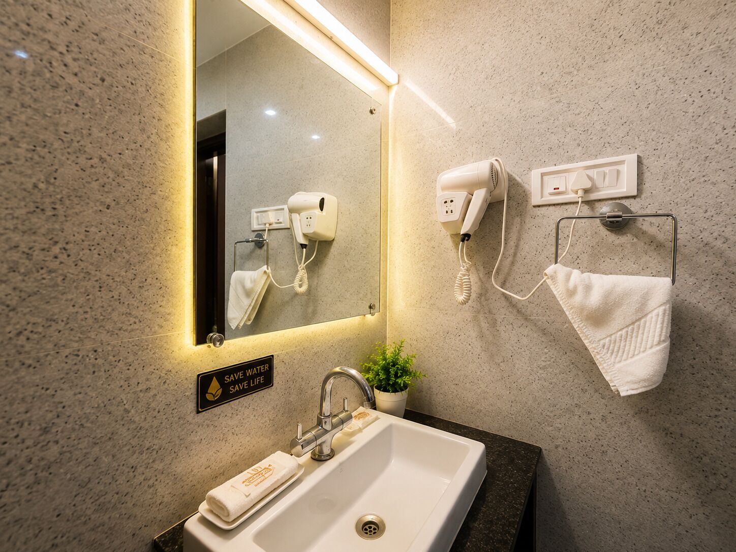 Un baño con lavamanos, espejo, toallero, secador de pelo y un cartel que dice 'SAVE WATER SAVE LIFE'.