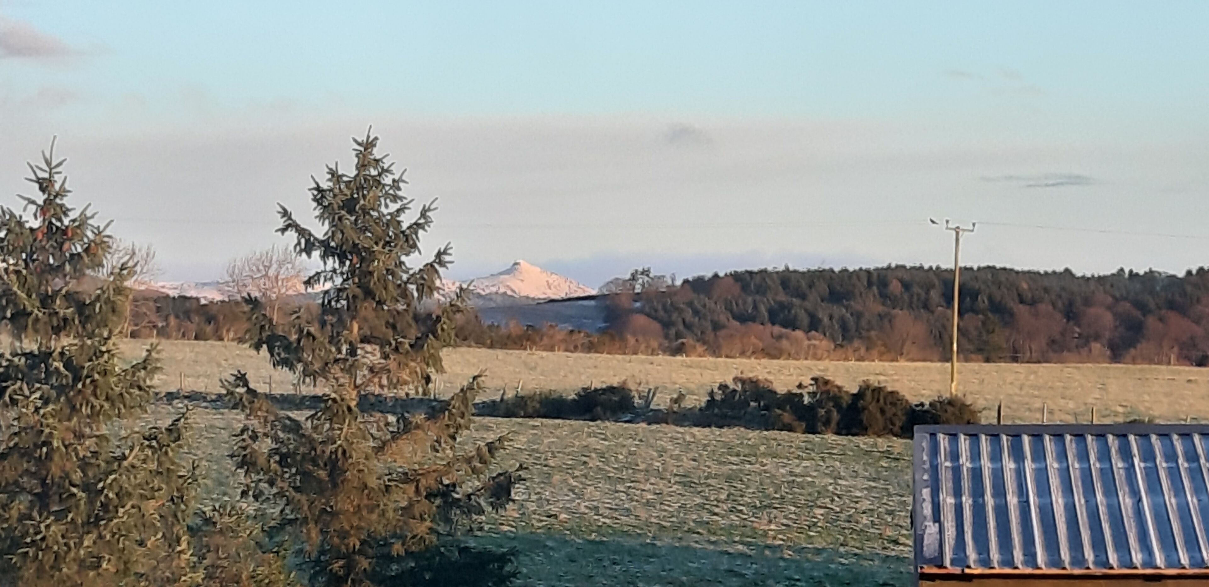 Paisaje rural con una montaña al fondo, un campo y un edificio con techo de láminas de metal.
