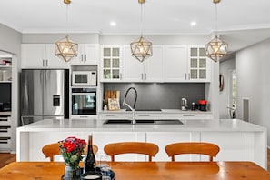 A modern kitchen with white cabinets, a stainless steel refrigerator, a built-in oven, and a large island with a sink. There are three pendant lights above the island. - Saltwater (Anglesea)