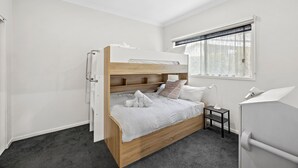 Ein Etagenbett mit Holzrahmen und weißer Bettwäsche, ein kleiner Nachttisch mit Lampe, ein Fenster mit Spitzenvorhängen und ein weißer Kleiderschrank.